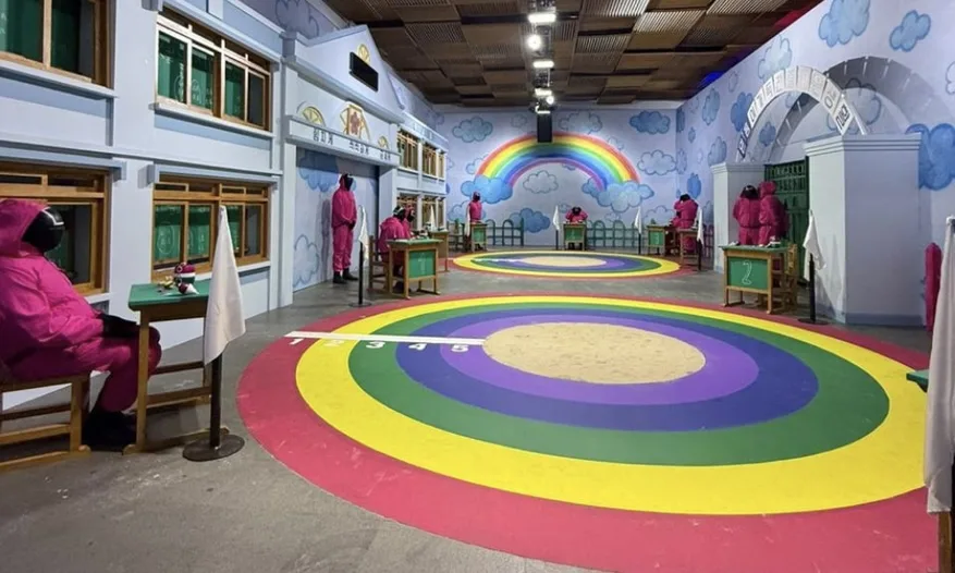 SQUID GAME — The classroom: marbles on the painted floor and the guards in pink