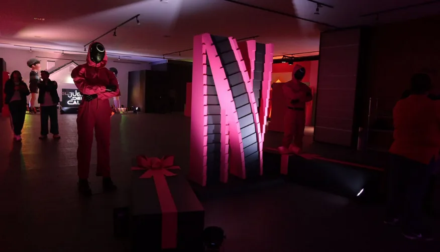SQUID GAME — Inside: the neon N, the guards in pink and the gift box